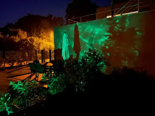 Gästegarten bei Nacht mit grün angestrahlter Hauswand, davor Büsche und ein Sitzplatz mit geschlossenem Sonnenschirm