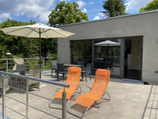 Dachterrasse mit zwei orangefarbenen Liegen und Sonnenschirm vor der Glasfront des Wohnbereichs