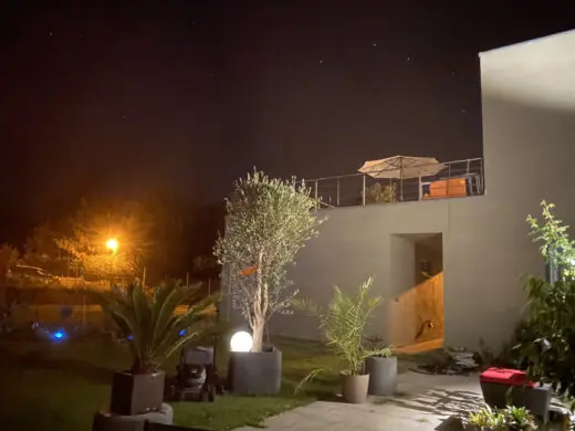 Hauswand mit Dachterrasse bei Nacht, davor beleuchteter Garten mit Olivenbaum und Kugelleuchte